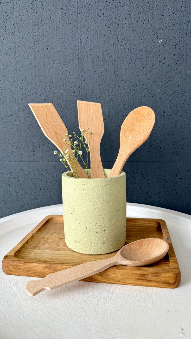 Pu&ograve; includere: Un set di utensili da cucina in legno, tra cui un cucchiaio, una spatola e un cucchiaio pi&ugrave; piccolo, sono esposti in un portautensili in cemento verde chiaro. Il portautensili &egrave; appoggiato su un vassoio di legno.