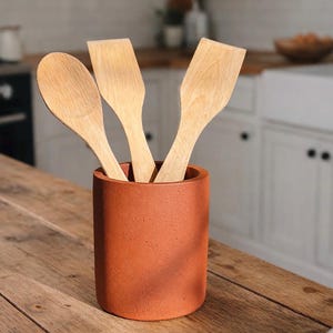 Puede incluir: Un portautensilios cilíndrico de color terracota con tres utensilios de cocina de madera. Los utensilios de madera incluyen una cuchara y dos espátulas. El soporte está sobre una superficie de madera, con una cocina al fondo.