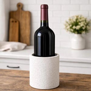 Puede incluir: Una botella llena de vino tinto en un enfriador de vino blanco texturizado. La botella de vino tiene una etiqueta roja oscura y una parte superior granate. El enfriador es cilíndrico y se encuentra sobre una superficie de madera.