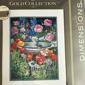 May include: A cross-stitch kit featuring a detailed garden scene. The image showcases a birdbath surrounded by colorful flowers, including roses, poppies, and daisies. A blue bird and a butterfly are also visible. The kit is from The Gold Collection by Dimensions.