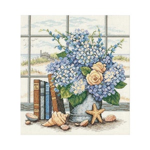 Könnte beinhalten: Ein Kreuzstichmuster mit einem Metalleimer voller blauer Hortensien und blassgelber Rosen, platziert neben drei Büchern und mehreren Muscheln. Die Szene ist vor einem Fenster mit Blick auf einen Sandstrand und Dünen dargestellt.