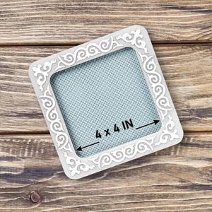 Puede incluir: Un marco cuadrado blanco con un diseño ornamental y arremolinado. El marco tiene una abertura de 10,16 x 10,16 cm con un fondo azul claro y punteado. El marco está sobre una superficie de madera.
