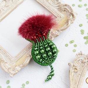 Puede incluir: Broche artesanal con forma de cardo, con una parte superior de plumas burdeos y un cuerpo de cuentas verdes. El broche está adornado con cuentas verdes y detalles rojos, creando un diseño floral detallado. El broche está colocado sobre un marco decorativo.