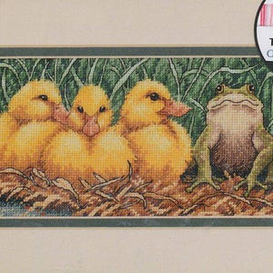 May include: Cross-stitch artwork featuring three yellow ducklings and a green frog. The ducklings are nestled together in a nest of brown leaves and grass, while the frog sits upright. The artwork is framed with a green border. The text "Pre-Sorted Thread Organizer" is visible.
