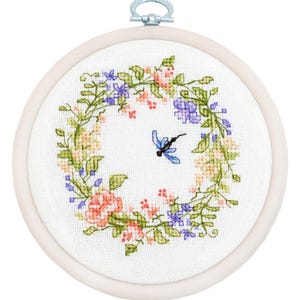May include: A cross-stitch embroidery hoop featuring a floral wreath design with pink, purple, and yellow flowers, and green leaves. A blue dragonfly is in the center. The embroidery is set in a round wooden hoop with a metal clasp.