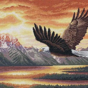 May include: A cross-stitch landscape depicts a bald eagle in flight against a sunset. The eagle has brown wings and a white head and tail. Mountains and a lake are in the background, with a warm orange and yellow sky.