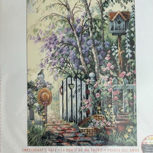 May include: A cross-stitch kit featuring a detailed garden scene with a white gate, brick pathway, and a birdhouse. The kit includes pre-sorted thread, 14-count ivory Aida fabric, a needle, and instructions. Finished size is 10 x 14 inches.
