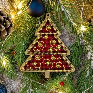 May include: A Christmas tree ornament with a red and gold beaded design. The tree is shaped with a wooden outline and features a detailed pattern. The ornament is surrounded by pine needles, lights, and other festive decorations.