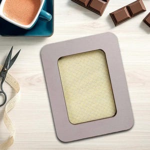 May include: A rectangular, light gray needlepoint frame with a beige mesh interior. The frame is surrounded by a light wood surface, with a blue cup of coffee, scissors, ribbon, and chocolate bars.