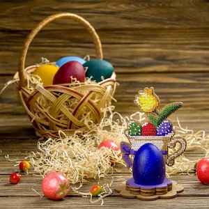 May include: A wooden Easter egg holder with a blue egg, a yellow flower, and a basket of colorful eggs in the background.