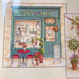 May include: A counted cross stitch pattern titled Afternoon in Provence featuring a teal storefront with the word VINTAGE above a window display of a chandelier, books, and a birdcage. A pink table and chairs sit outside with potted plants.
