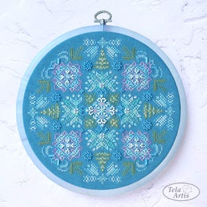 Puede incluir: Un bastidor de bordado de punto de cruz azul, con un diseño detallado de copo de nieve. El diseño incorpora tonos de azul, blanco y toques de rosa y verde. El bordado está en un bastidor azul claro, creando una pieza decorativa.