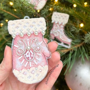 Könnte beinhalten: Rosa-weißer, mit Perlen besetzter Weihnachtsbaumschmuck in Form eines Fäustlings mit Schneeflockenmuster. Der Fäustling ist mit Perlen und Kristallen verziert. Ein zweiter Schmuck im Hintergrund ist ein rosa Eislaufschuh. Die Ornamente hängen an einem Weihnachtsbaum.