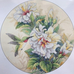 May include: A cross stitch pattern featuring a hummingbird hovering near a cluster of white hibiscus flowers with green leaves. The pattern is designed by Lena Liu.