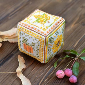 May include: A cube-shaped, hand-stitched needlepoint featuring autumn-themed designs. The cube showcases a sunflower, pumpkins, and a maple leaf, all in shades of yellow, orange, and green. The background is a light gray, with a brown border.