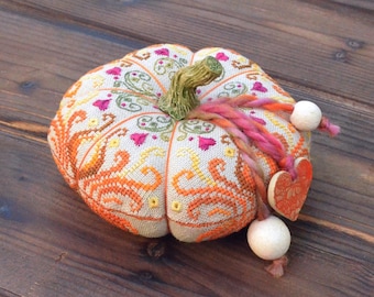 May include: A decorative, handcrafted pumpkin with intricate cross-stitch embroidery in orange, yellow, pink, and green. It features a green stem, a braided yarn accent, and wooden heart and ball embellishments. The pumpkin is a fall-themed decorative item.