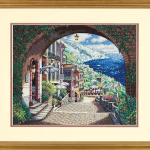 Puede incluir: Obra de arte en punto de cruz enmarcada que representa una escena de un pueblo costero italiano. La obra presenta una calle adoquinada que conduce al mar, enmarcada por un arco. Se ven edificios con balcones y sombrillas. El marco es dorado.