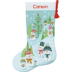 May include: A light blue Christmas stocking with a winter scene featuring snowmen, a Christmas tree, and falling snowflakes. The word "Carson" is printed in red at the top. A red tassel hangs from the top.