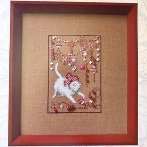 May include: Cross-stitch artwork of a white cat with a red bow, playing with a ball. The artwork is framed in a red wooden frame. The background is a tan fabric with red and white decorative elements.