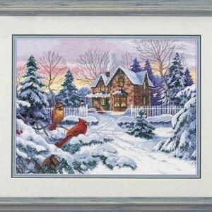 May include: A framed cross-stitch embroidery depicting a winter scene with a stone house, snow-covered pine trees, and two red cardinals perched on a branch in the foreground. The artwork is enclosed in a light blue-grey wooden frame with a white mat.