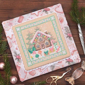 May include: A cross-stitch of a gingerbread house with a pink roof, surrounded by Christmas trees and gifts. The cross-stitch is framed with a green and white border and set on a patterned fabric with Christmas cookies. A pair of gold scissors and a glass ornament are in the foreground.
