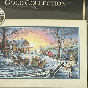 May include: A Dimensions Gold Collection cross-stitch kit featuring a winter scene. The image depicts a horse-drawn sleigh carrying people through a snowy landscape, with a house, church, and sunset in the background. The kit includes a thread organizer.