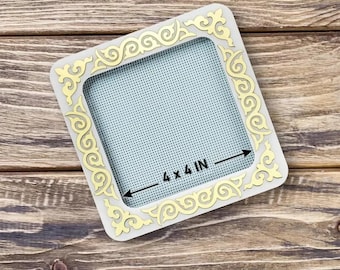 May include: Square, off-white frame with a gold filigree design. The frame has a 4 x 4 inch opening with a light blue mesh backing. The frame is on a wooden surface.