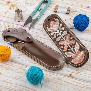 Puede incluir: Un estuche de cuero marrón para tijeras y un estuche a juego con un diseño floral de punto de cruz. También se muestran tijeras turquesas, un dedal y ovillos de lana amarilla y azul, lo que sugiere un tema artesanal. Los estuches miden unos 15 cm.