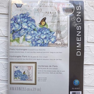 May include: A counted cross stitch kit featuring a hydrangea flower with a butterfly and the Eiffel Tower in the background. The kit includes presorted thread, 14 count ivory Aida fabric, a needle, and instructions. The design is by Julie Paton. The kit is 14 inches by 11 inches or 35.5 centimeters by 27.9 centimeters.