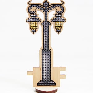 May include: A laser-cut wooden ornament shaped like a double-headed street lamp. The design features intricate cross-stitch detailing in shades of grey and black, with yellow thread accents highlighting the two lantern heads. It stands on a base.