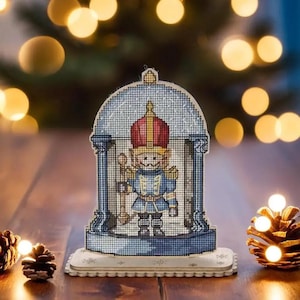 May include: A wooden Christmas ornament featuring a Nutcracker soldier in a blue uniform with a red hat and gold trim. The ornament is decorated with a white snow-like design and the words "The Nutcracker" are printed on the top.