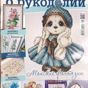 Puede incluir: Una portada de revista con un patrón de punto de cruz de un lindo conejito que lleva un vestido azul y un ribete de piel blanca. El título de la revista es "Все о рукоделии" y el texto "Майские фантазии" está debajo del conejito. La portada de la revista también presenta otros patrones de punto de cruz de flores, una casa y un bolso.
