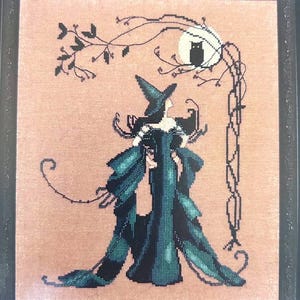 May include: Cross-stitch art of a witch in a teal gown and hat, framed in black. A black owl sits on a branch under a full moon. The witch is standing under a tree branch with leaves.