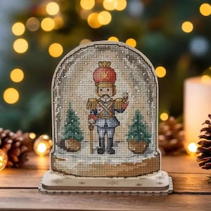May include: A decorative snow globe featuring a cross-stitch design of a nutcracker soldier. The soldier wears a red hat and blue uniform, standing between two small green Christmas trees. The globe sits on a wooden base, perfect for holiday decor.