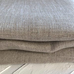 Può includere: Primo piano di tessuto di lino piegato, color naturale. Il tessuto ha una trama grossa e una superficie leggermente strutturata. Le pieghe creano ombre e profondità, evidenziando la drappeggiatura e il peso del tessuto. Il colore è beige neutro.