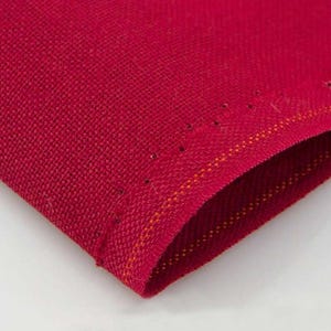 May include: Close-up of a section of red fabric with a textured weave. The edge is folded over, revealing a contrasting orange thread. Small, evenly spaced holes are visible along the fold, adding a decorative element to the textile.
