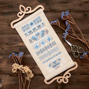 May include: A cross-stitch sampler featuring various blue floral and geometric patterns. The sampler is displayed on a wooden scroll holder. Dried flowers and decorative scissors are also present, creating a rustic aesthetic.