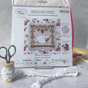 May include: A cross-stitch embroidery kit featuring a lamb in strawberries design. The kit includes a printed fabric, seed beads, and fluffy thread. The package also includes a pair of scissors and a wooden spool with white lace.