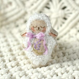 May include: A small stuffed sheep ornament featuring a cross-stitched face with closed eyes and pink cheeks, surrounded by white textured faux fur. The sheep has a light purple ribbon tied in a bow at the neck and a purple floral embroidery design.