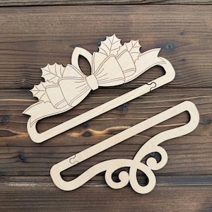 Op de afbeelding: Twee houten kleerhangers. Eén heeft een strik- en hulstbladontwerp, de andere heeft een decoratieve krul. Beide zijn gemaakt van lichtgekleurd hout en staan tegen een donkere houten achtergrond.