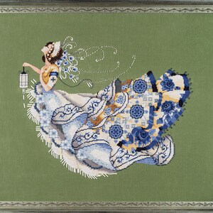 Può includere: Opera d'arte a punto croce di una donna in un abito fluente, che tiene in mano una lanterna. L'abito presenta intricati motivi floreali blu e bianchi con accenti dorati. Lo sfondo è verde unito e l'opera è incorniciata.