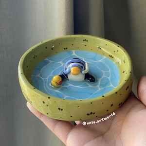 Puede incluir: Un cuenco de cerámica con exterior amarillo salpicado de puntos oscuros. En el interior, un diseño de agua azul y blanco presenta un pingüino con snorkel y un pequeño pato amarillo. El cuenco se sostiene en una mano.