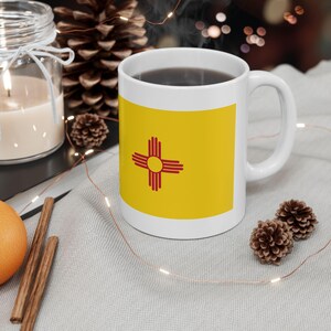 May include: White ceramic coffee mug with a yellow and red design of the New Mexico state flag. The mug is filled with coffee and sitting on a white surface.