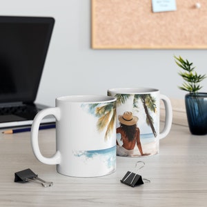 May include: Two white ceramic mugs with a beach scene printed on them. The scene features a woman in a straw hat sitting on a beach with palm trees in the background.