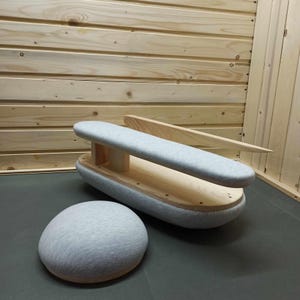 Op de afbeelding: Een houten kleermakerspersham met een lichtgrijze gewatteerde oppervlakte en een apart rond perskussen. De ham heeft een gebogen houten basis en een houten arm. De achtergrond is een licht houten paneel.