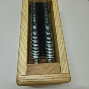 May include: A rectangular light-coloured wooden frame housing two parallel rows of stacked dark metal washers. The metal components are secured with silver-coloured bolts and nuts at the base, creating a structured, industrial-style assembly.