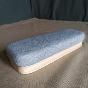 Op de afbeelding: Een strijkplankhoes van grijs stof met een houten basis. Het hoes is rechthoekig en heeft een afgeronde rand. De basis is van licht hout.