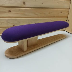 Op de afbeelding: Een mouwstrijkplank met een lang, cilindervormig, met paarse stof bekleed kussen op een lichtgekleurde basis van natuurlijk hout. De structuur bestaat uit een afgerond bovendeel dat wordt ondersteund door een centraal houten blok.