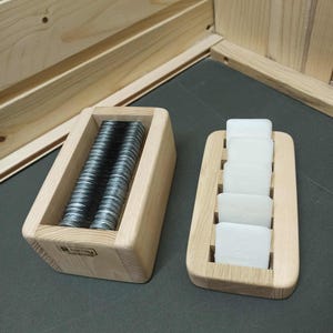 May include: Two wooden trays. One contains a stack of silver metal bolts. The other holds five white rectangular soap bars. The trays are made of light-colored wood and are set against a dark gray surface.