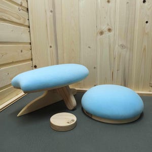 Op de afbeelding: Twee lichtblauwe meditatiekussens, een met een houten basis en drie poten, de andere met een ronde houten basis. Beide kussens zijn gemaakt van zachte stof.